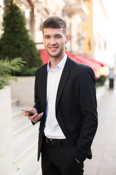 Young beautiful smiling man in black suit and white shirt with wireless earphones holding cellphone in hand happily looking in camera spending time on street