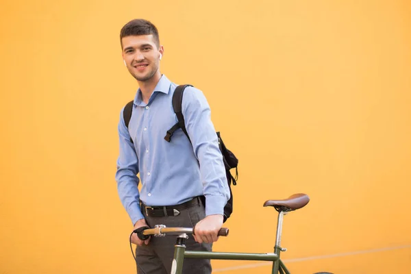 Young attractive man in blue shirt and backpack with wireless earphones standing with retro bicycle dreamily looking in camera over orange background