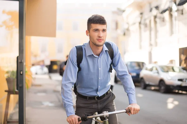 Young attractive man in blue shirt with wireless earphones riding bicycle thoughtfully looking aside with cozy street on background