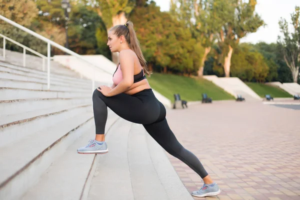 Young beautiful plus size woman in sport top and leggings dreamily stretching on stairs in city park