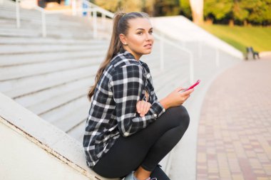 Young beautiful plus size woman in casual shirt and leggings leaning on stair railing thoughtfully looking aside with cellphone in hand spending time in park clipart