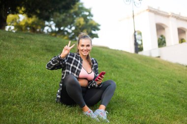 Young pretty smiling woman in casual shirt and leggings holding cellphone in hand happily showing two fingers gesture spending time on grass in park