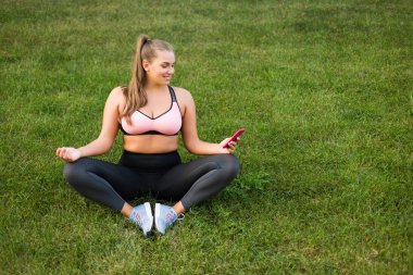 Genç, gülümseyen, uzun boylu, pembe spor bluzlu ve taytlı bir kadın. Parktaki yeşil çimlerde meditasyon yapan bir cep telefonu kullanıyor.