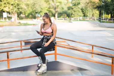 Genç gülümseyen güzel artı boyutu kadın pembe sportif üst, tozluk ve mutlu bir şekilde paten parkta cep telefonu kullanarak korkuluk üzerine eğilerek paten 