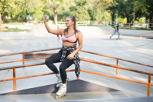 Genç gülümseyen güzel artı boyutu kadın pembe sportif üst, tozluk ve mutlu bir şekilde paten parkta vakit cep telefonu üzerinde fotoğraf çekme korkuluk üzerine eğilerek paten 