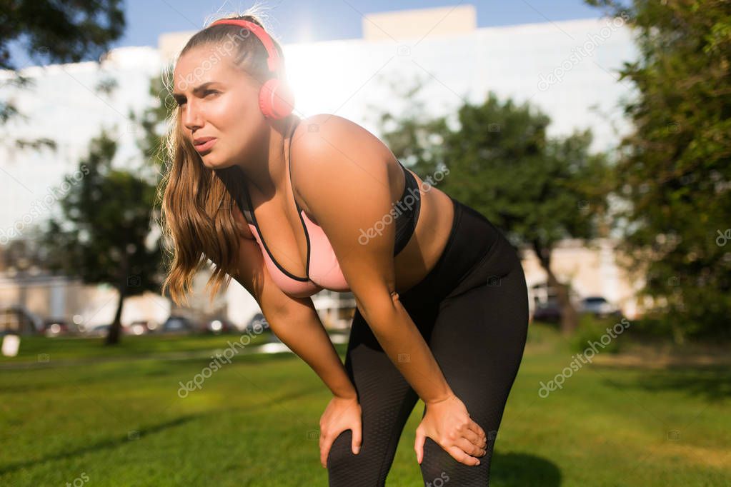Young tired plus size woman in sporty top and leggings with red headphones thoughtfully looking aside leaning on knees in city park 