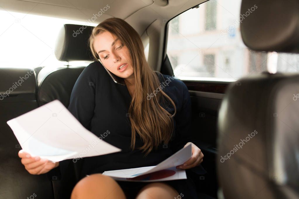 Young busy businesswoman in black dress sitting on back seat in car thoughtfully talking on cellphone with documents in hands