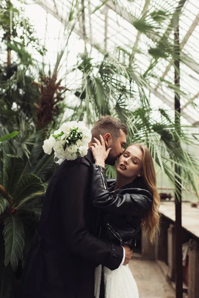 Young beautiful woman in black leather jacket and white dress with little bouquet of flowers in hand dreamily closing eyes attractive man embracing and kissing her in check in cozy greenhouse