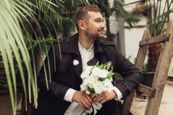 Young handsome smiling man in black trench coat and shirt holding little bouquet of white flowers in hands dreamily looking aside leaning on old wood ladder in beautiful cozy greenhouse