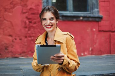 Turuncu trençkot ve tablet mutlu kamera arıyorum eski avluda elinde tutan siyah puantiyeli elbise genç çekici neşeli kadın