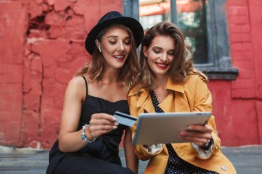 Kredi kartı turuncu trençkot mutlulukla birlikte eski avluda vakit tablet holding neşeli kadınla elinde tutan radyo earphones ile siyah şapkalı kadın genç güzel