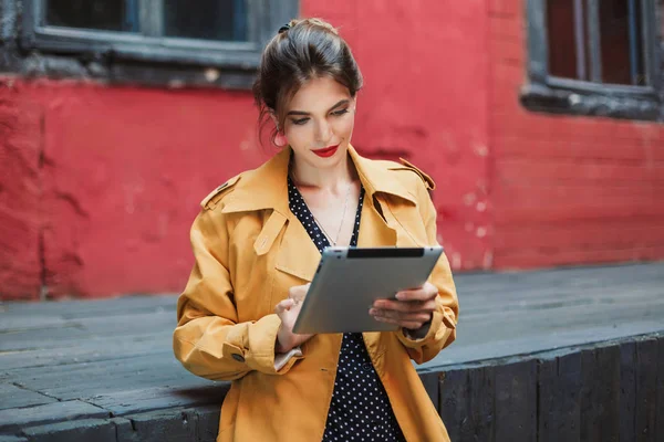 年轻迷人的女人在橙色风衣和黑色波尔卡点礼服若有所思地使用平板电脑花时间在老院子里 — 图库照片