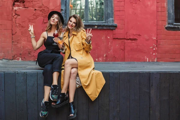 Two young beautiful women with cocktails in hands happily looking in camera together showing two fingers gestures spending time in old cozy courtyard of cafe