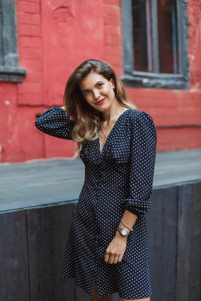 Young attractive woman in black polka dot dress with red lips happily looking in camera spending time in old courtyard