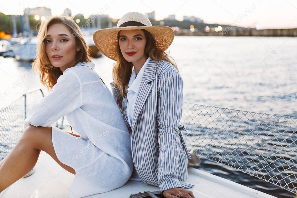 Young beautiful woman in white shirt and gorgeous lady in stripped jacket and straw hat dreamily looking in camera together on yacht with amazing harbor view on background