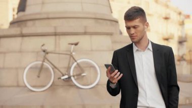Genç kasvetli iş adamı ciddiye cep telefonu ekrana bakarak ve bir bisiklet arka plan ve kablosuz kulaklık ile bir Şehir Meydanı genelinde swiping siyah takım elbise