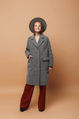 Gri ceket ve geniş şapka ile çekici genç gülümseyen kadın sersem kapanış gözleri bej renkli arka plan üzerinde pantolon.