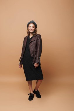 Oldukça şapkalı kız gülümseyerek, deri ceket ve siyah elbise sevinçle kamera bej renkli arka plan üzerinde seyir