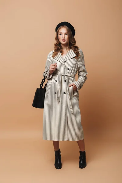 Young attractive woman with wavy hair in striped coat and black hat holding bag in hand dreamily looking aside over beige background