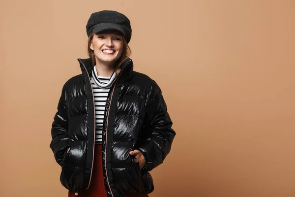 Young beautiful smiling woman with wavy hair in cap and black down jacket joyfully looking aside over beige background