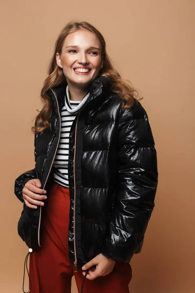 Young cheerful woman with wavy hair in black down jacket happily looking aside over beige background
