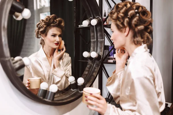 Young beautiful actress with wavy hairstyle and red lips in silk robe applying powder dreamily looking in mirror spending time in dressing room
