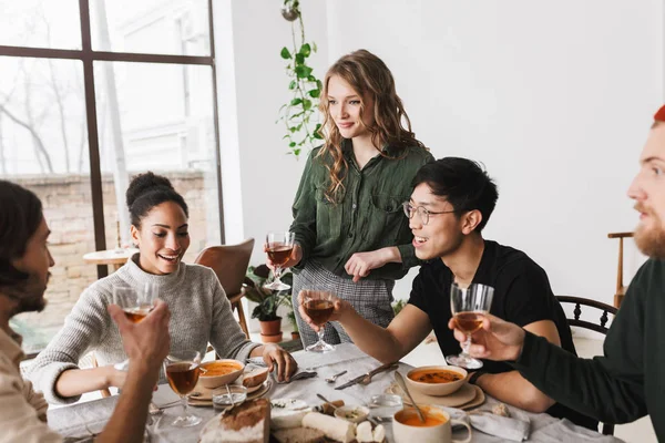 Beautiful woman with wavy hair holding glass of wine in hand standing near table full of food. Group of attractive  international friends happily spending time together having lunch in cozy cafe