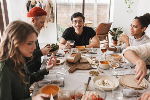 Радостный азиат в очках с бокалом вина в руке, счастливо смотрящий в камеру. Группа привлекательных международных друзей, сидящих за столом и обедающих вместе в уютном кафе

