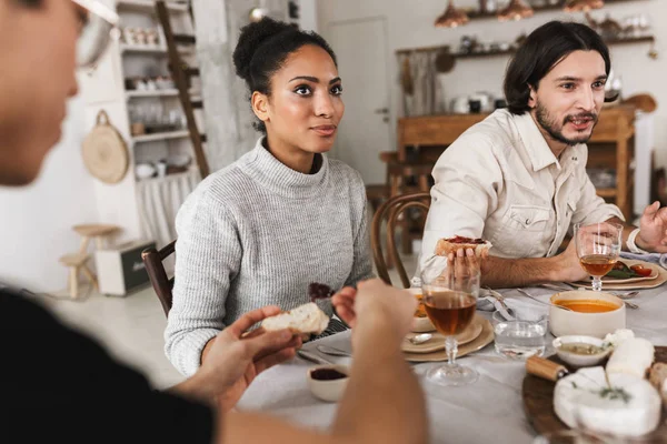 Dreamy african american woman with dark curly hair sitting at the table thoughtfully looking aside. Group of young international friends spending time together on lunch in cozy cafe