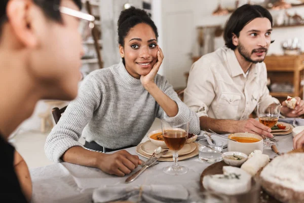 Smiling african american woman with dark curly hair sitting at the table leaning on hand thoughtfully looking man. Group of young international friends spending time together on lunch in cozy cafe