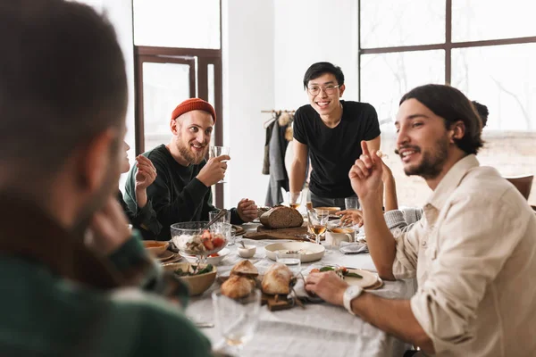 Young cheerful asian man in eyeglasses and black T-shirt leaning on table joyfully looking aside. Group of attractive international friends happily spending time together on lunch in cozy cafe
