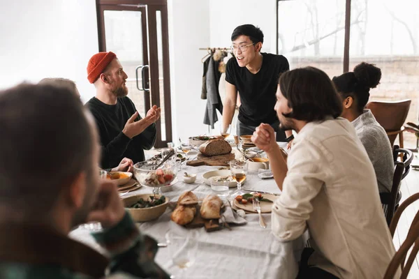 Young asian man in eyeglasses and black T-shirt leaning on table happily talking with colleagues. Group of attractive international friends spending time together on lunch in cozy white cafe