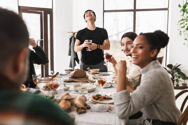 Young laughing asian man in eyeglasses and black T-shirt happily holding glass of wine in hand. Group of attractive international friends joyfully spending time together on lunch in cozy cafe