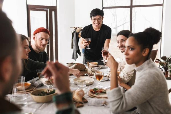 Young smiling asian man in eyeglasses and black T-shirt happily holding glass of wine in hand. Group of attractive positive international friends spending time together on lunch in cozy cafe