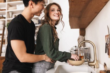 Çok gülümseyen kadın mutlu bir kenara çamaşır bulaşık Asya adam onu kucaklayan arkasında seyir. Sevinçle birlikte evde rahat mutfakta vakit genç uluslararası çift