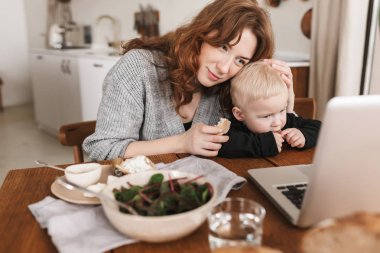Örme kazak gıda ile masada oturan ve laptop çizgi film izlerken küçük oğlu sarılma Kızıl saçlı genç güzel kadın. Anne ile bebek evde rahat mutfakta vakit