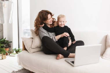 Kamera mutlu seyir o küçük gülümseyen oğlu ile kanepede oturan genç güzel kadın. Anne ve bebek çocuk sevinçle dizüstü evde rahat oturma odasında ile vakit