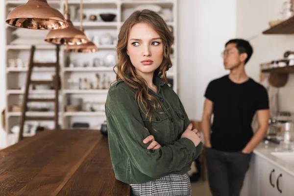 Young beautiful woman with wavy hair holding hands together sadly looking aside with asian man on background. International couple quarrel spending time on kitchen at home