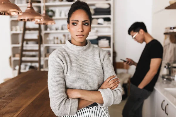 Upset african american woman holding hands together thoughtfully looking aside with asian man holding cellphone on background. Young international couple in quarrel spending time on kitchen at home