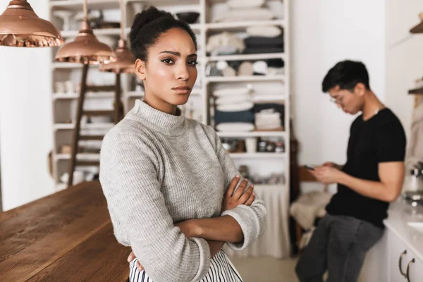 Upset african american woman holding hands together sadly looking in camera with asian man holding cellphone on background. Young international couple in quarrel spending time on kitchen at home