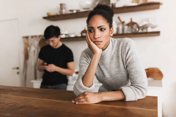 Thoughtful african american woman leaning on hand sadly looking aside with asian man using cellphonr on background. Young international couple in quarrel spending time on kitchen at home