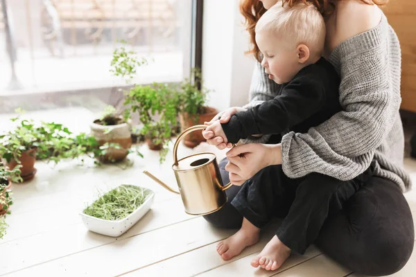 Young beautiful woman sitting on floor with her little son holding watering can in hands with green plants around near big window. Mom dreamily spending time with baby boy at cozy home