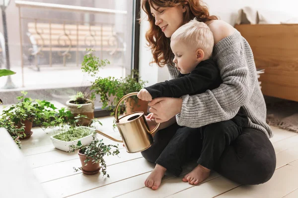 Young beautiful smiling woman sitting on floor with her little son holding watering can in hands with green plants around near big window. Mom happily spending time with baby boy at cozy home