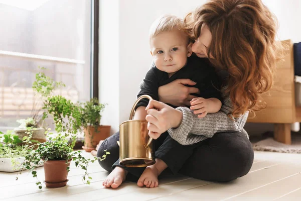 Young attractive woman sitting on floor hugging her little handsome son holding watering can in hands with green plants around near big window. Mom happily spending time with baby boy at cozy home