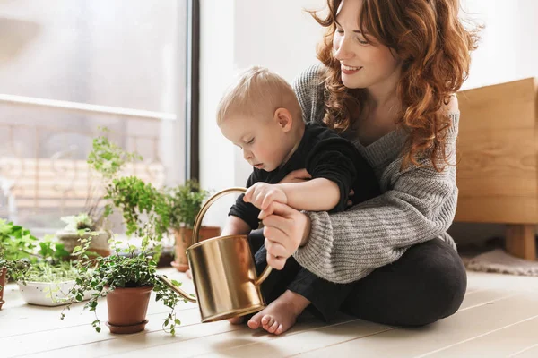 Young smiling woman sitting on floor with her little handsome son holding watering can in hands with green plants around near big window. Mom joyfully spending time with baby boy at cozy home