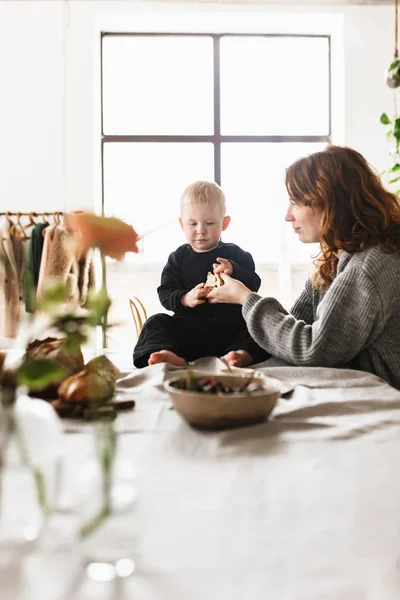 Young beautiful mom with red hair in knitted sweater dreamily playing with her little handsome son spending time together in cozy kitchen at home