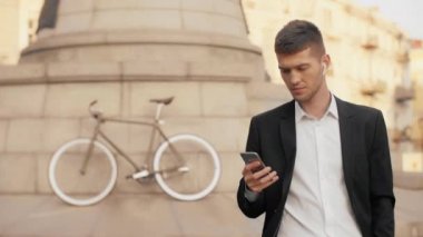 Genç ciddi iş adamı dikkatle cep telefonu ekrana bakarak ve bir bisiklet arka plan ve kablosuz kulaklık ile bir Şehir Meydanı genelinde swiping siyah takım elbise