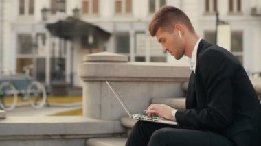 Genç adam üstünde belgili tanımlık kucak tutan dizüstü bilgisayarda yazarak Suite. Bisiklet arka planda ile Şehir Meydanı'nda oturan işyeri dışında tipik kablosuz kulaklık ile işadamı konsantre
