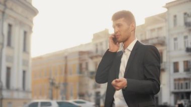 Gün batımında güneş parlama ile Kent Meydanı, telefonda konuşurken iş adamı. Siyah Suite cep telefonu sokak sıcak sabah güneş ışığında el hareketi ile konuşurken genç adam