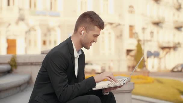 套房里的年轻人手里拿着笔记本电脑打字 微笑的商人带着无线耳机在办公室外打字 坐在城市广场的楼梯上 — 图库视频影像